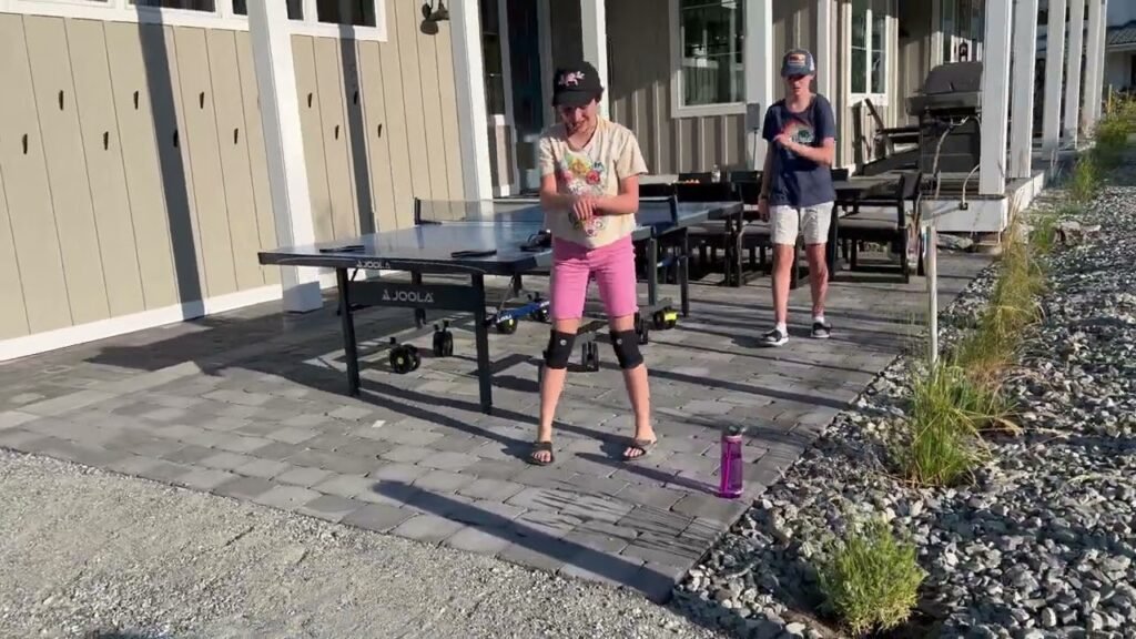 Two friends enjoying an outdoor game of table tennis on a sunny deck with a gravel path and greenery.