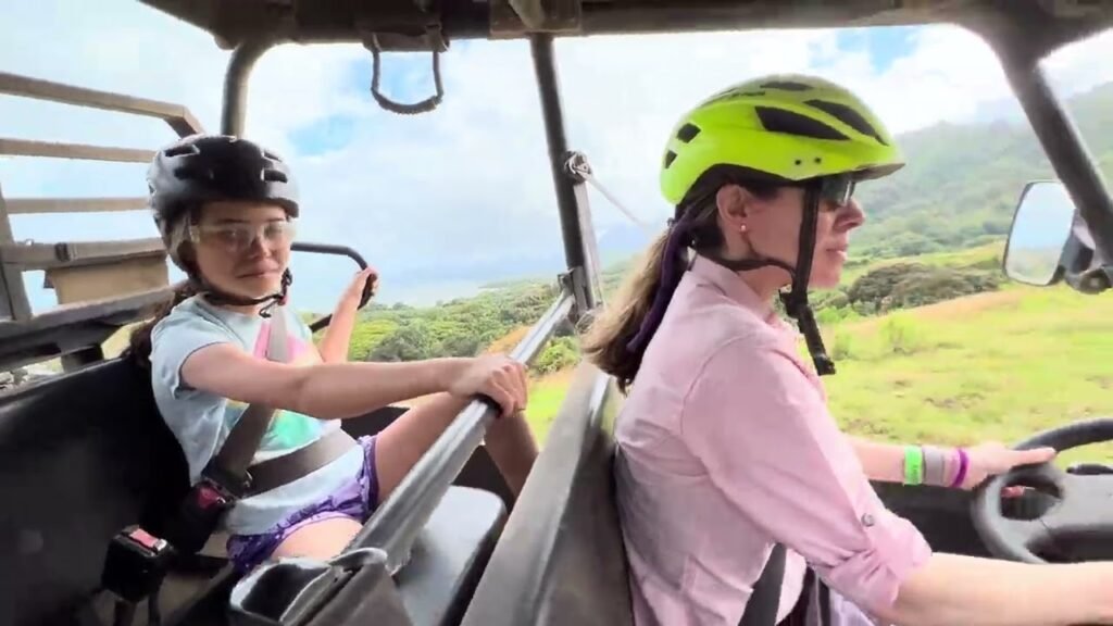 Two people wearing helmets enjoy a ride in a safari truck with a scenic landscape in the background.