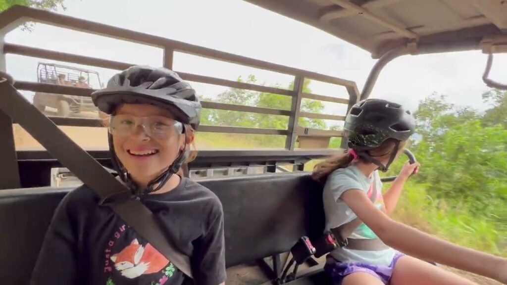 Children wearing helmets on an open vehicle during a journey. They are secured with seatbelts and enjoying the outback environment.