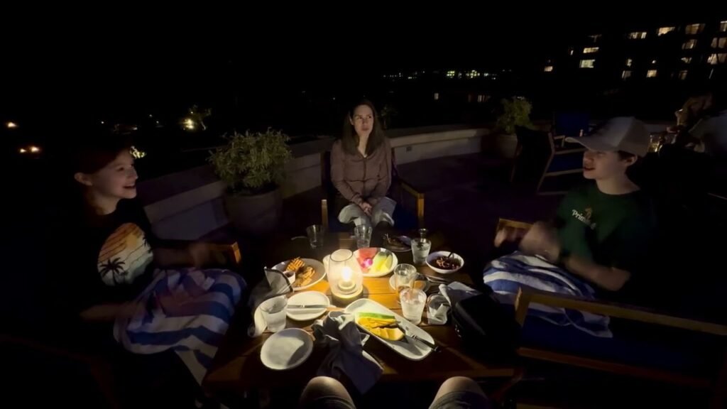 People enjoying a well-lit evening meal on a rooftop dining table overlooking distant lights in the dark.