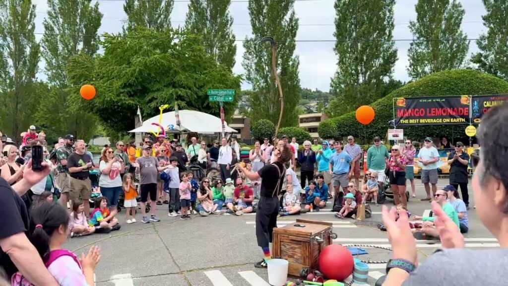 A lively crowd gathers outdoors at the Village Lemonade to watch a juggler in the street.