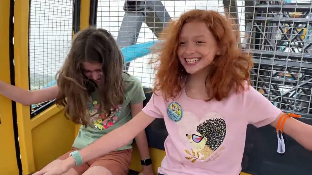 Two smiling visitors enjoy a ride on a yellow observation gondola above a busy station. Bright cables and a grid-like structure are visible through the safety screens.