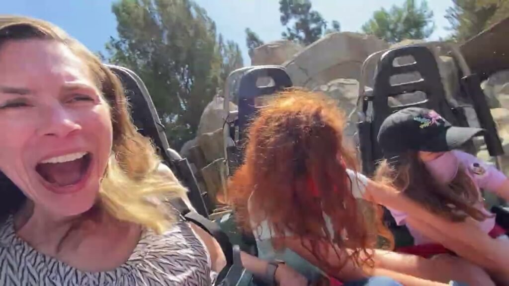 A woman screams with excitement while riding a roller coaster. Children are visible in the seats behind her.