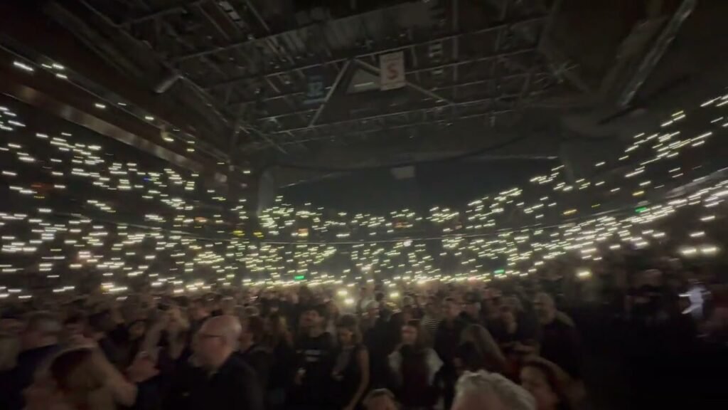 Audience holding up illuminated phones creating a glowing field under the venue ceiling during a large-scale performance.