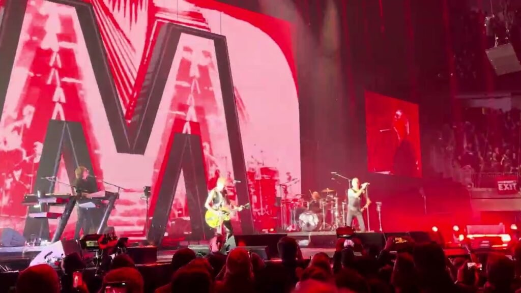 A band performs on stage under a massive glowing letter M with red lighting at a live concert, as the audience captures the moment on their phones.