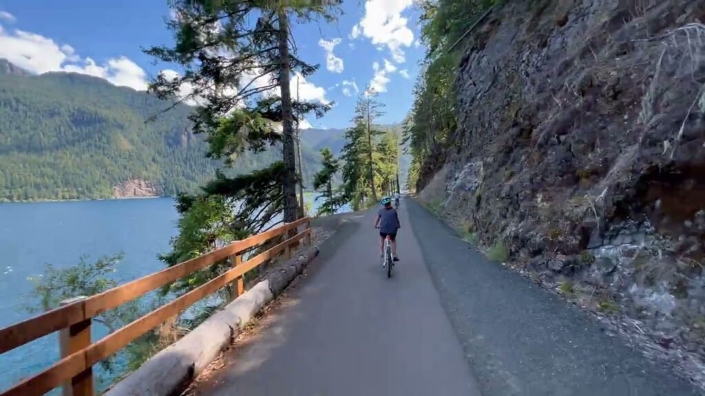 A scenic paved path winds along a rocky cliffside with a view of blue water and distant mountains. A person is seen cycling away from the camera on the trail.