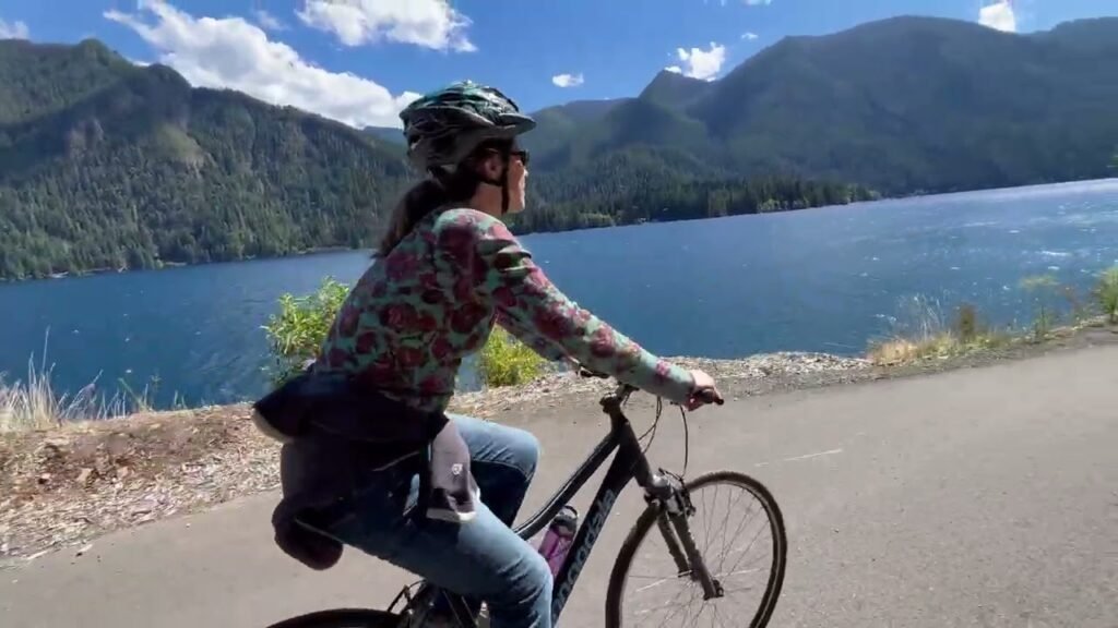 A person wearing a helmet rides a bike along a scenic road next to Lake Wenatchee, with the Osceola Dam visible in the distance.