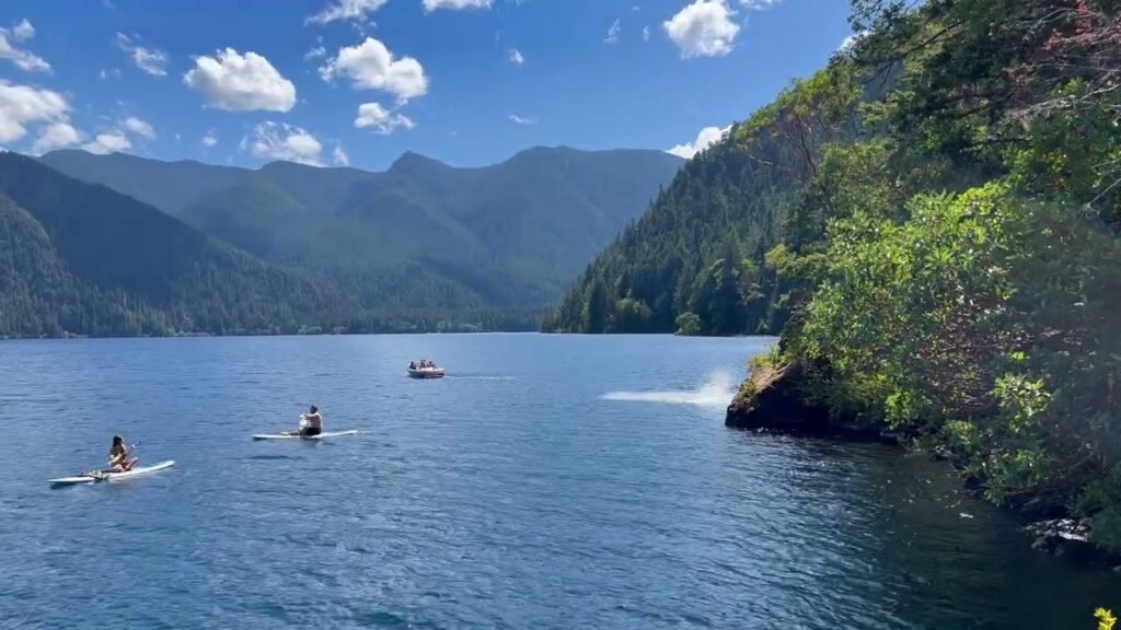 Kayakers paddle on a blue lake surrounded by forested mountains and a bright sky.