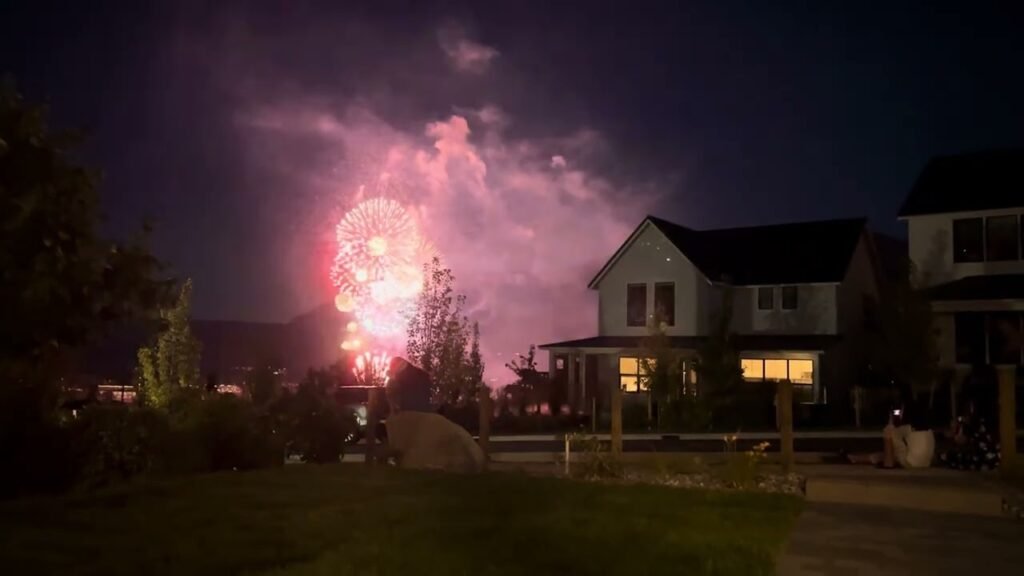 Vibrant pink fireworks illuminate the night sky above a residential neighborhood with houses and trees. Distant mountains are visible in the background under the glowing display.