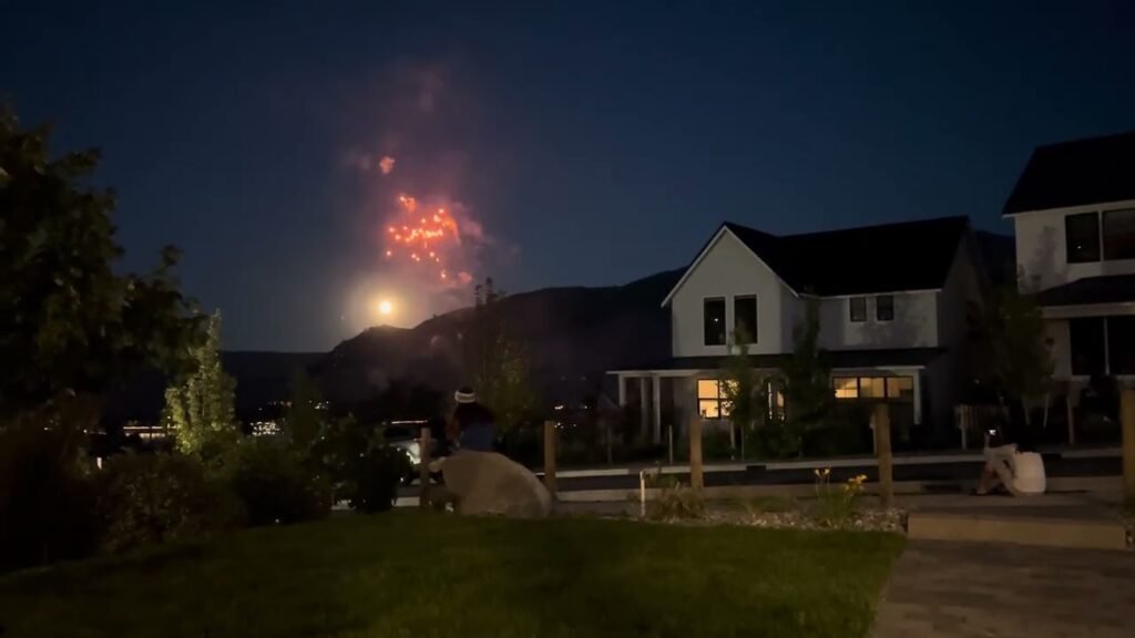 Fireworks light up the night sky above a suburban neighborhood, with houses and mountains silhouetted.