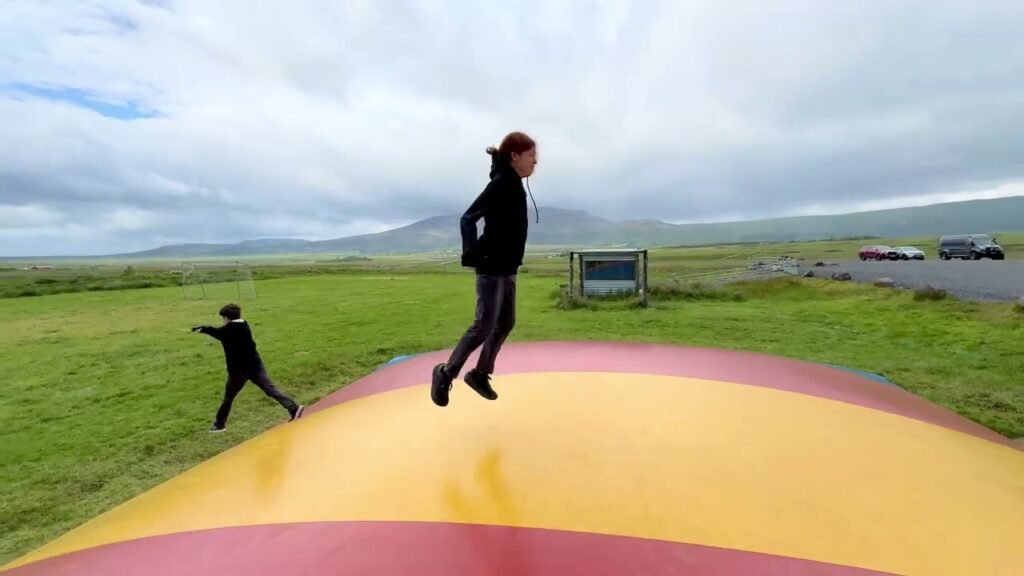 Two people bounce on a large, vibrant reindeer horn sculpture set against green Icelandic fields and distant mountains.