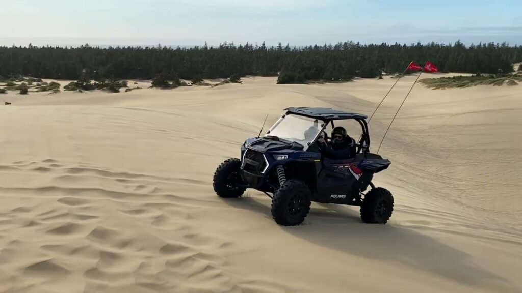 A side-by-side utility vehicle driving across Pacific Northwest sand dunes. A dense tree line marks the boundary with the surrounding forest.