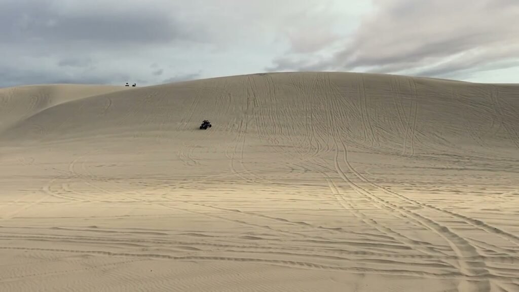 Vast desert sand dunes under a cloudy sky with a motor vehicle climbing the slope.