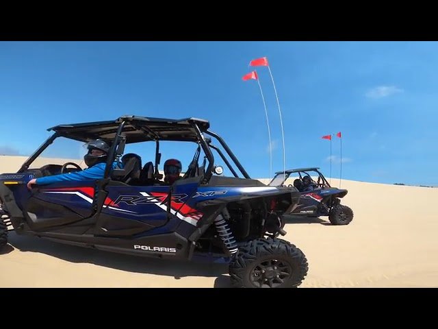 Polaris UTV vehicles racing across sand dunes under a clear blue sky. Red flags mark the course in the desert landscape.