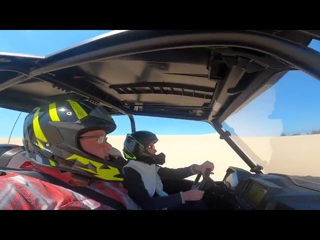Two helmeted riders navigate a dirt track from inside a racing vehicle under a sunny sky.
