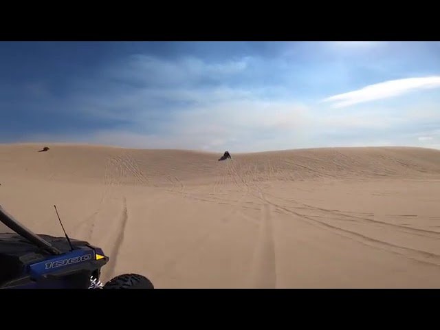 An off-road vehicle navigating vast sandy dunes under a dramatic sky. Tire tracks mark the path on the sand.
