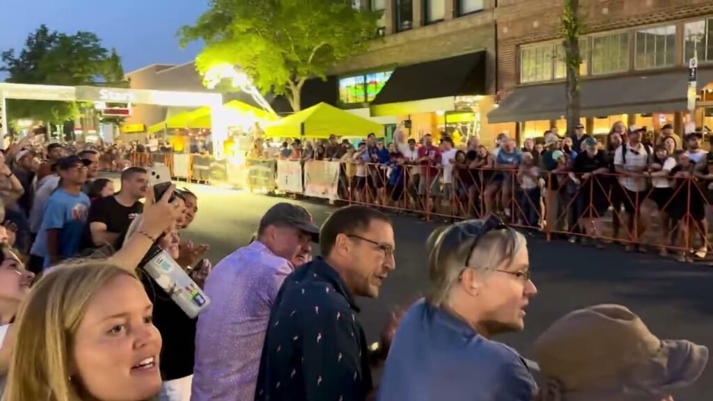 A large crowd gathers at an evening street market with yellow tents and brick buildings visible.