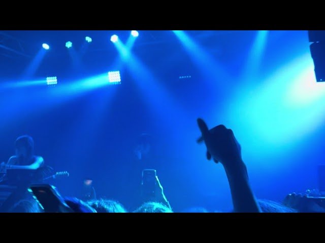 A dimly lit concert scene featuring blue stage lights, a crowd, and a silhouetted raised hand.