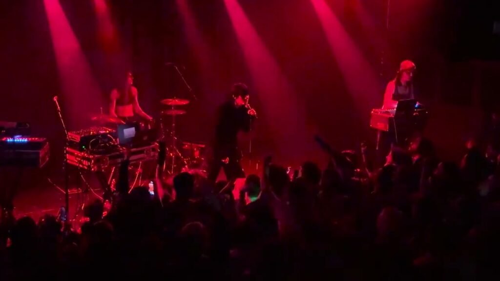 A band performs on stage under dancing red lights, with a packed crowd in the foreground.