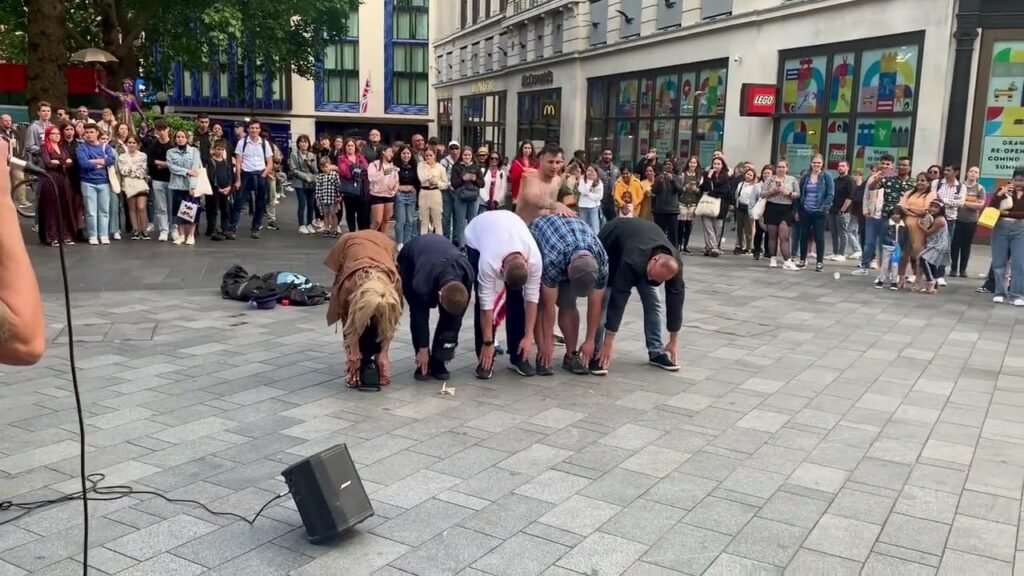 A group of performers bows to a crowd in a city plaza, with colorful storefronts in the background.
