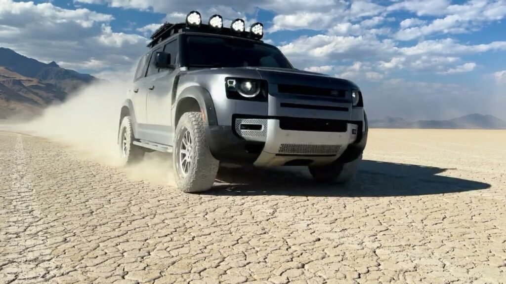 A gray Nissan Pathfinder is shown speeding over dry cracked desert ground. The scene includes vast sky in the distance with a dirt cloud behind the car.