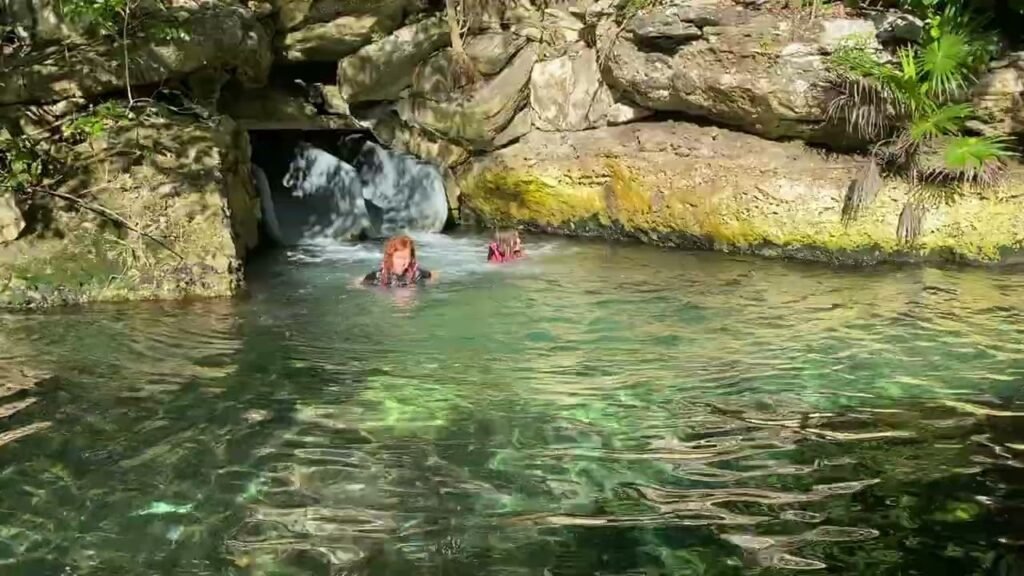 A person relaxing in a clear, natural pool beside a small waterfall and rocky cliff wall. Peaceful outdoor swimming scene with greenish-blue water.