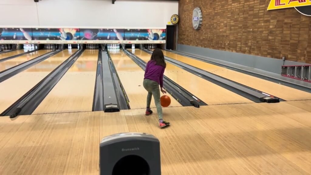 A person in a purple sweater prepares to roll an orange bowling ball down the lane. The background shows the bowling alley's pins and decor, highlighting a fun outdoor activity.