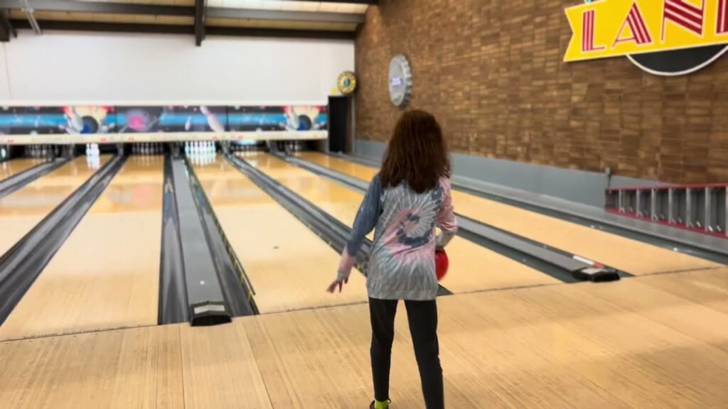 A person holds a bowling ball on the lane while aiming for the pins. The image features a classic bowling alley setup with a chalkboard background and "LANE" signage.
