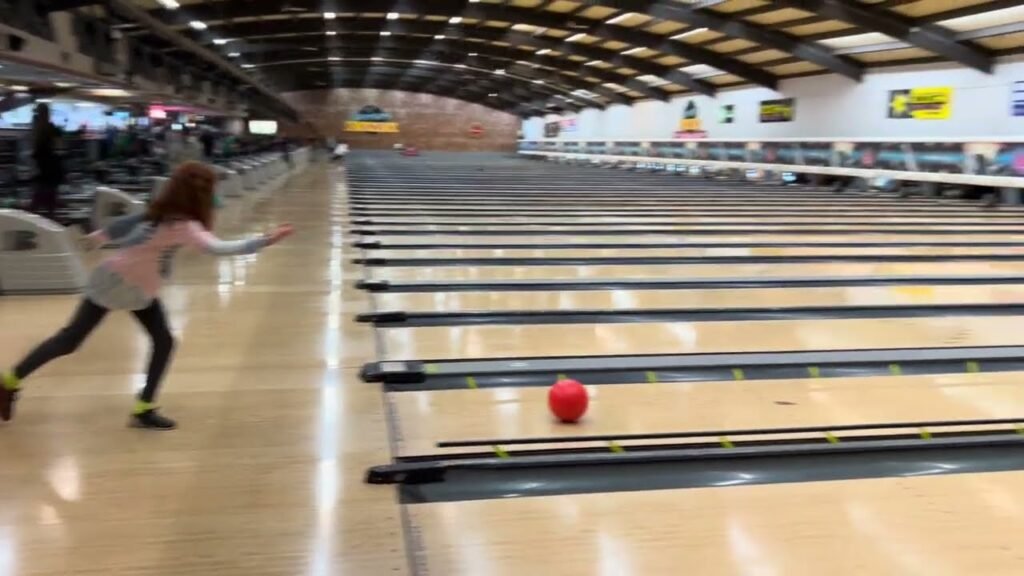 A person bowls at a brightly lit indoor facility, with a red ball rolling down the lane.