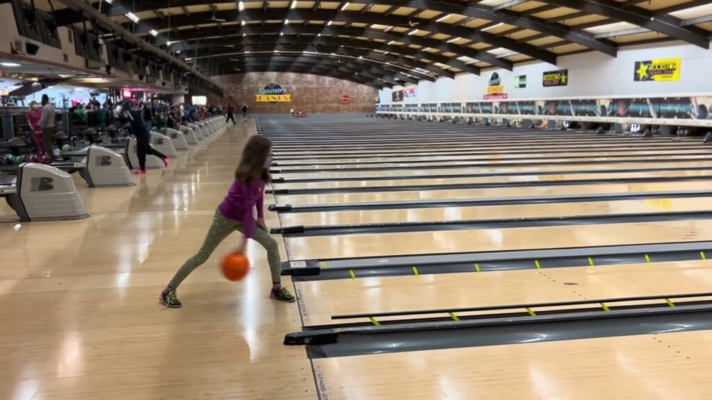 A person in a purple shirt holding an orange bowling ball in a bowling alley. The background shows multiple bowling lanes and seating.