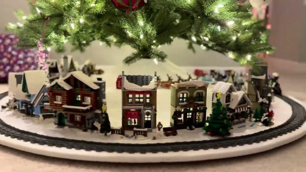 A festive Christmas tree skirt featuring a snow-covered village with miniature houses and trees. The decorative scene sits neatly under a lit Christmas tree.