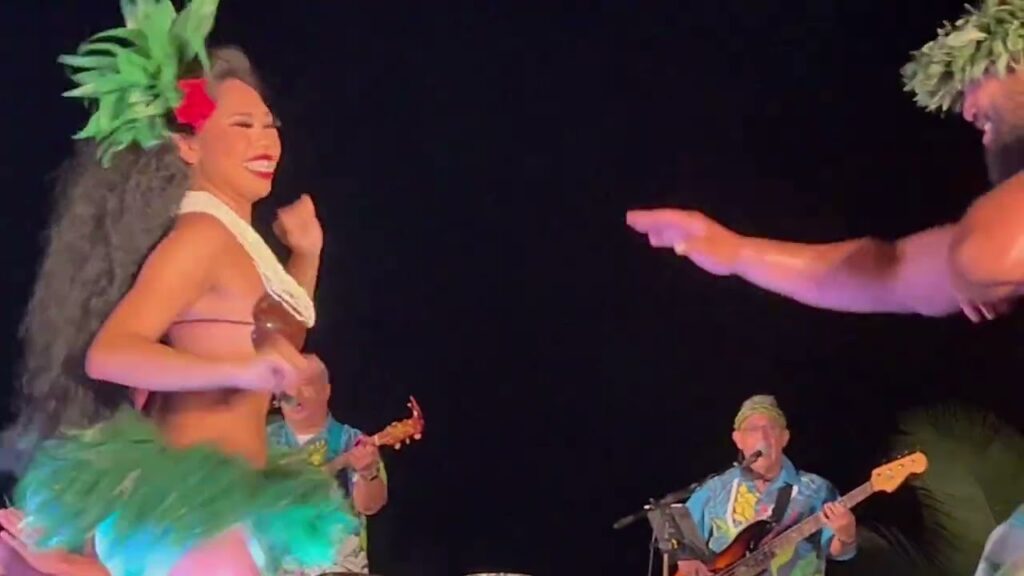 A live music stage featuring a dancer in traditional Polynesian attire and a band playing instruments under warm stage lighting.