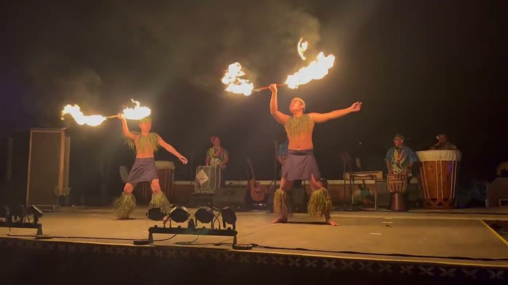 Fire dancers in vibrant purple pants and bare torsos swirling flaming poi on a concert stage. In the background, a band plays live with a large conga drum.