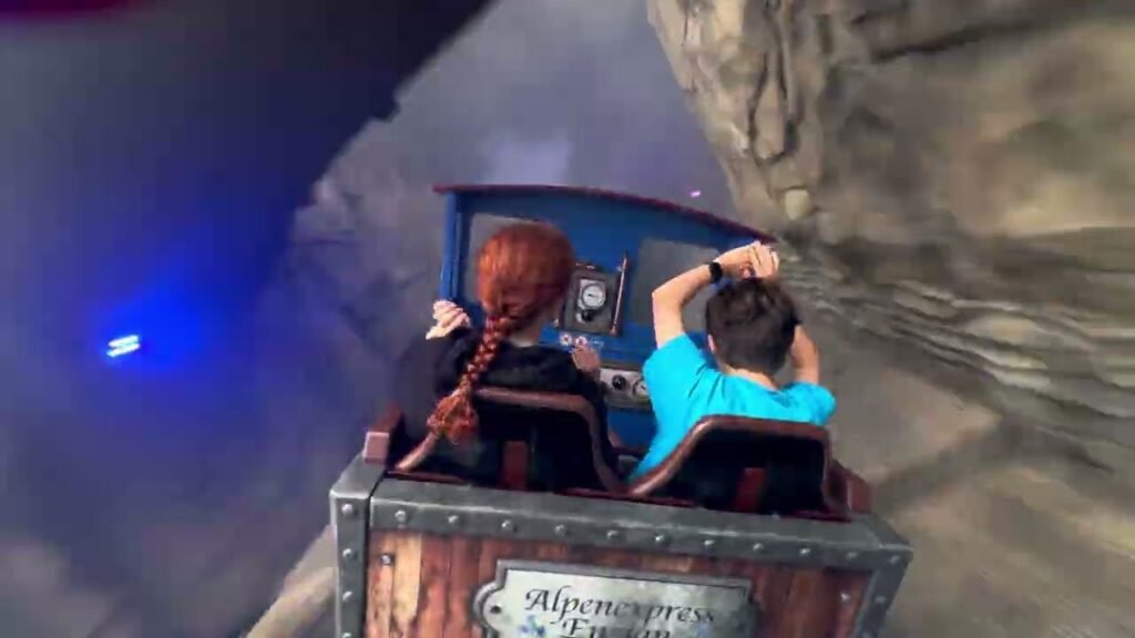 Two people enjoy a thrilling ride in the Alpenglow Express cart at a theme park, with one grinning in excitement and the other holding on tightly.