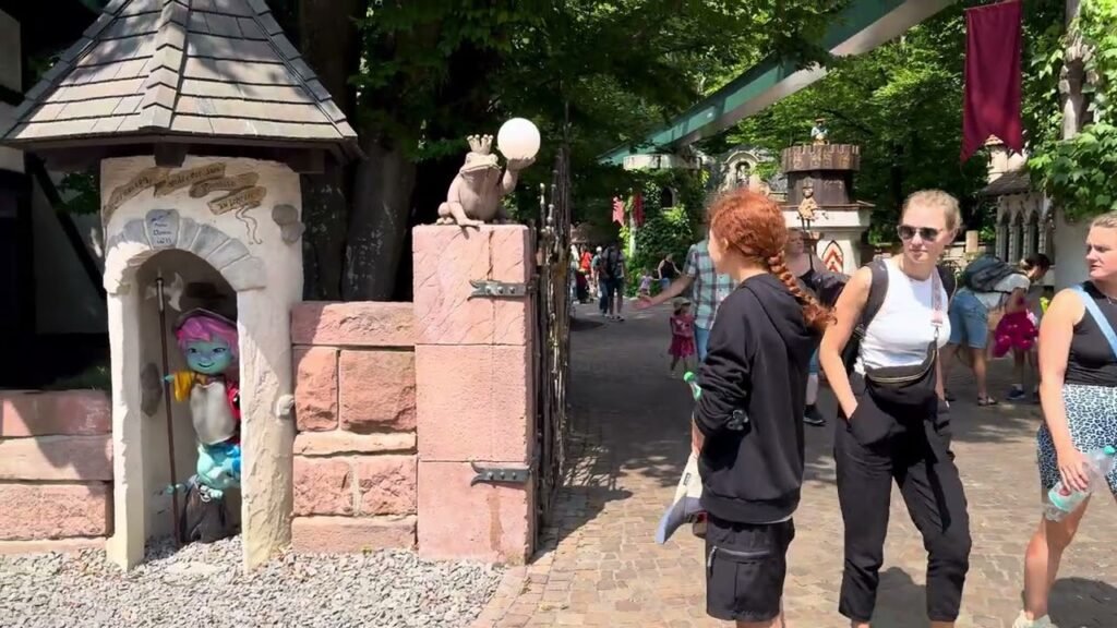 A sunny day in a picturesque village square with ornamental stone structures and pedestrians strolling.