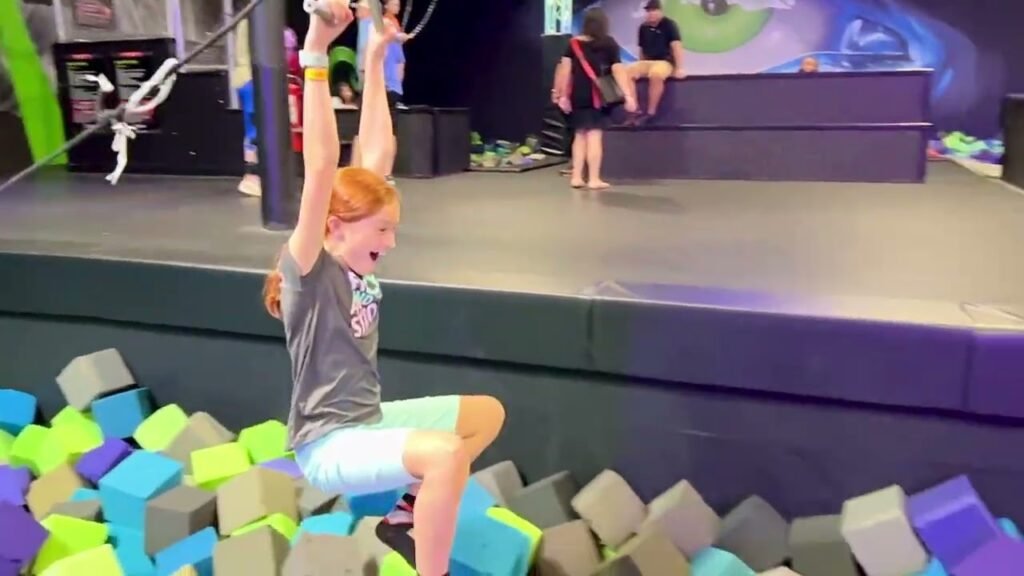 An individual hangs from a trampoline bungee cord above a foam pit at an indoor adventure park.
