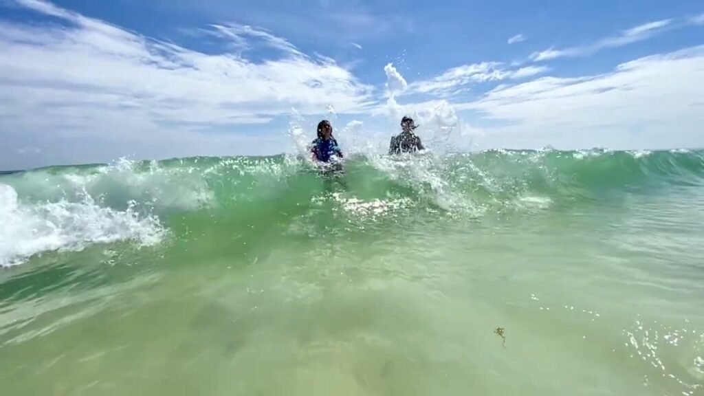 Two people paddleboarding on a deep green ocean wave under a partly cloudy blue sky.