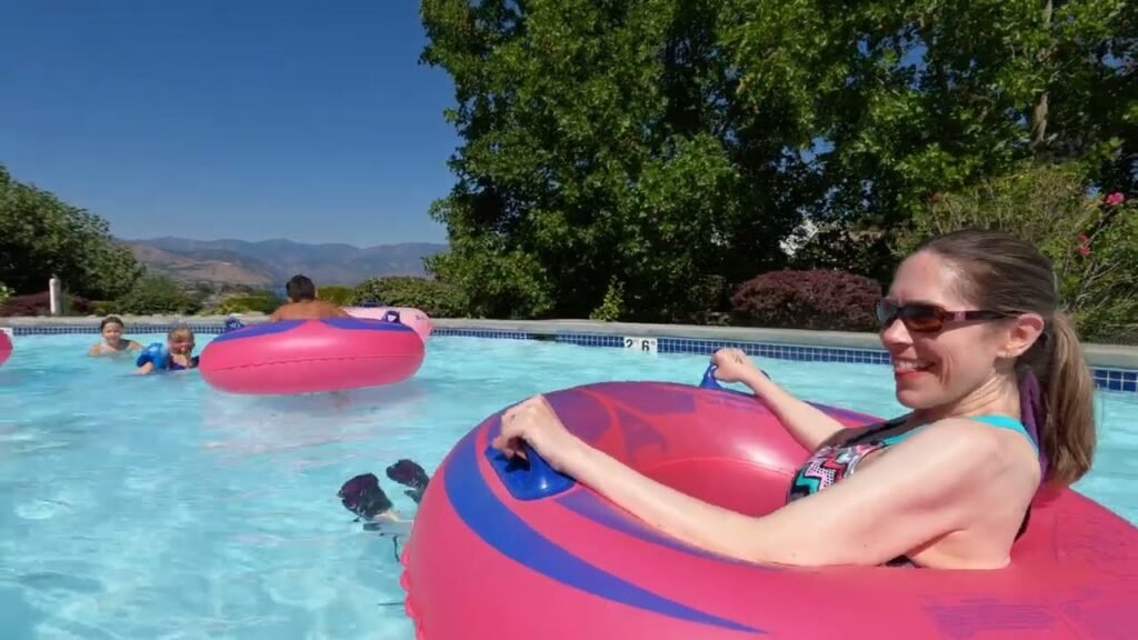 A person relaxes on a pink pool float in a sunny outdoor pool with a scenic mountain background.