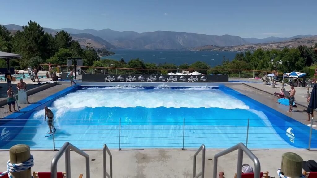 People enjoying a FlowRider surfing simulator next to a colorful pool with mountain views.