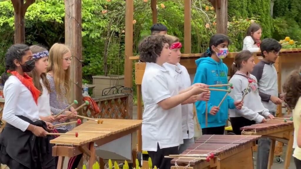 A group of young students and an instructor learn to play a wooden instrument in an outdoor forest setting. The participants are wearing safety eyewear and gathering around wooden tables.