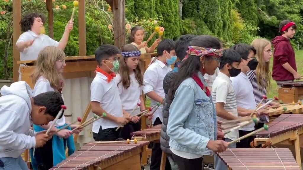 A group of campers participating in an outdoor marimba lesson on a sunny day. Participants are gathered around the instruments playing music in a forested area.