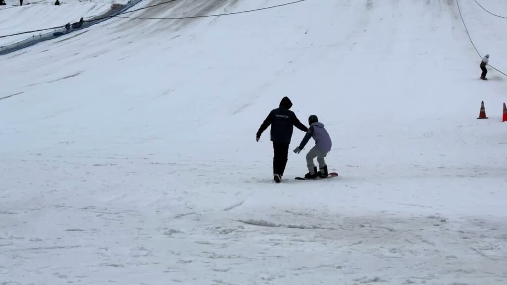 Two people on a ski slope, one helping the other balance on a snowboard.