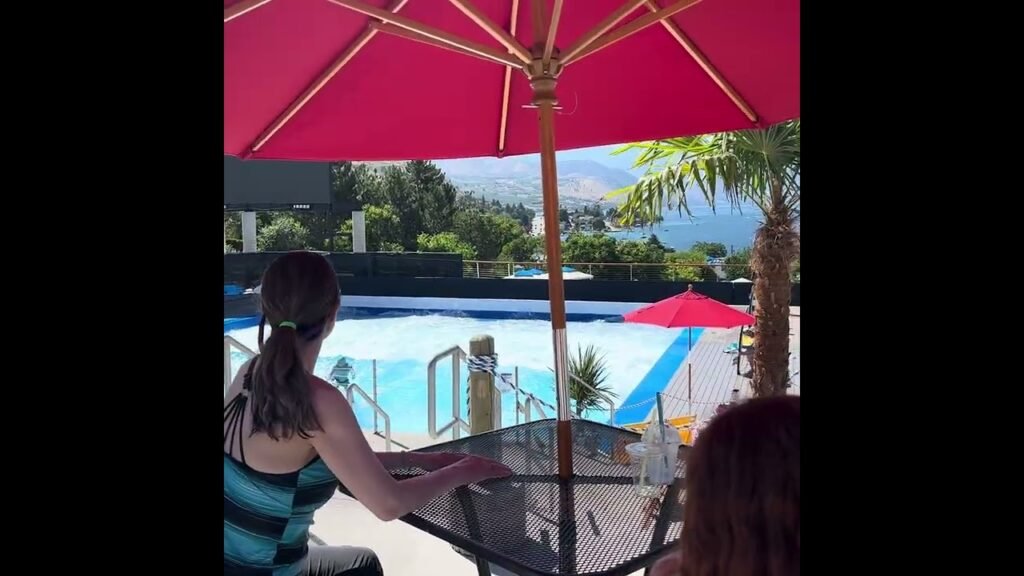 A view of a swimming pool with a blue slide from under a red patio umbrella with a person sitting at a table.