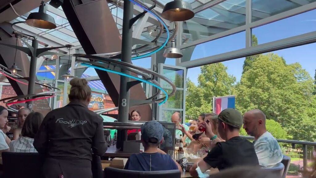 People enjoying a sunny afternoon at a modern rooftop cafe with large windows overlooking green trees and bright skies.