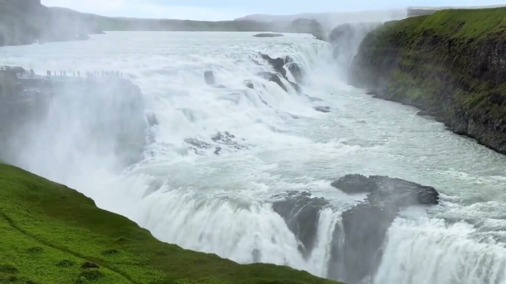 Majestic wide waterfall cascading into a river surrounded by mist and vibrant green hillside cliffs.