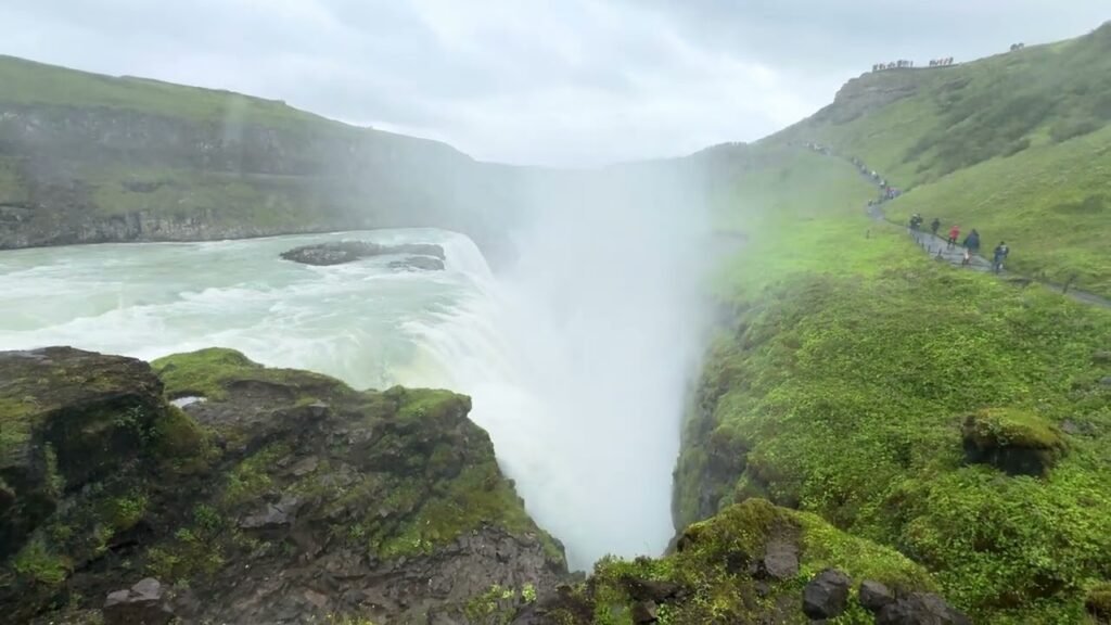 A dramatic waterfall cascading into a misty gorge with lush green hillsides and a hiking trail winding along the cliff edge.