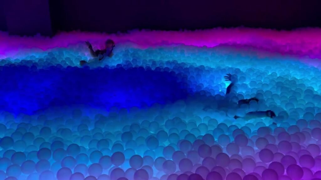 A person floats in a glowing ball pool illuminated with neon blue and pink lights at night.