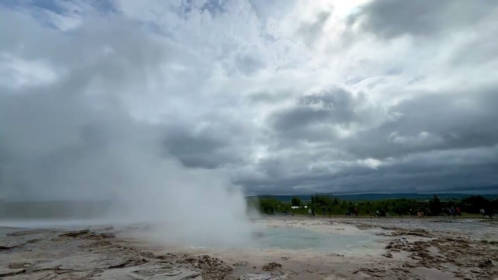 A large steam plume rises intensely from a hot spring under a heavy, overcast sky, with steam drifting over rugged terrain.