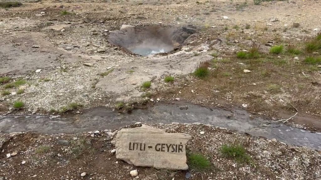 A stone sign labeled 'Li'Li - Geyser' sits in the foreground near a shallow stream and a geothermal basin in a desolate landscape.