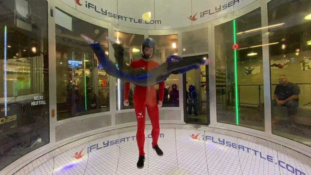 A person in a red flight suit stands beside a large glass wind tunnel at iFLY Seattle, ready for indoor skydiving.